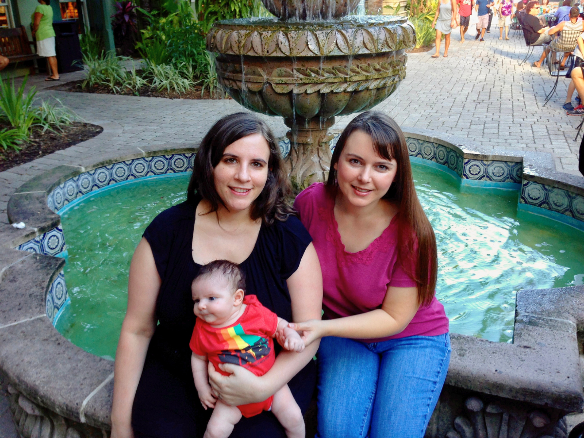 Two smiling women with a baby sit beside a decorative fountain in a courtyard.