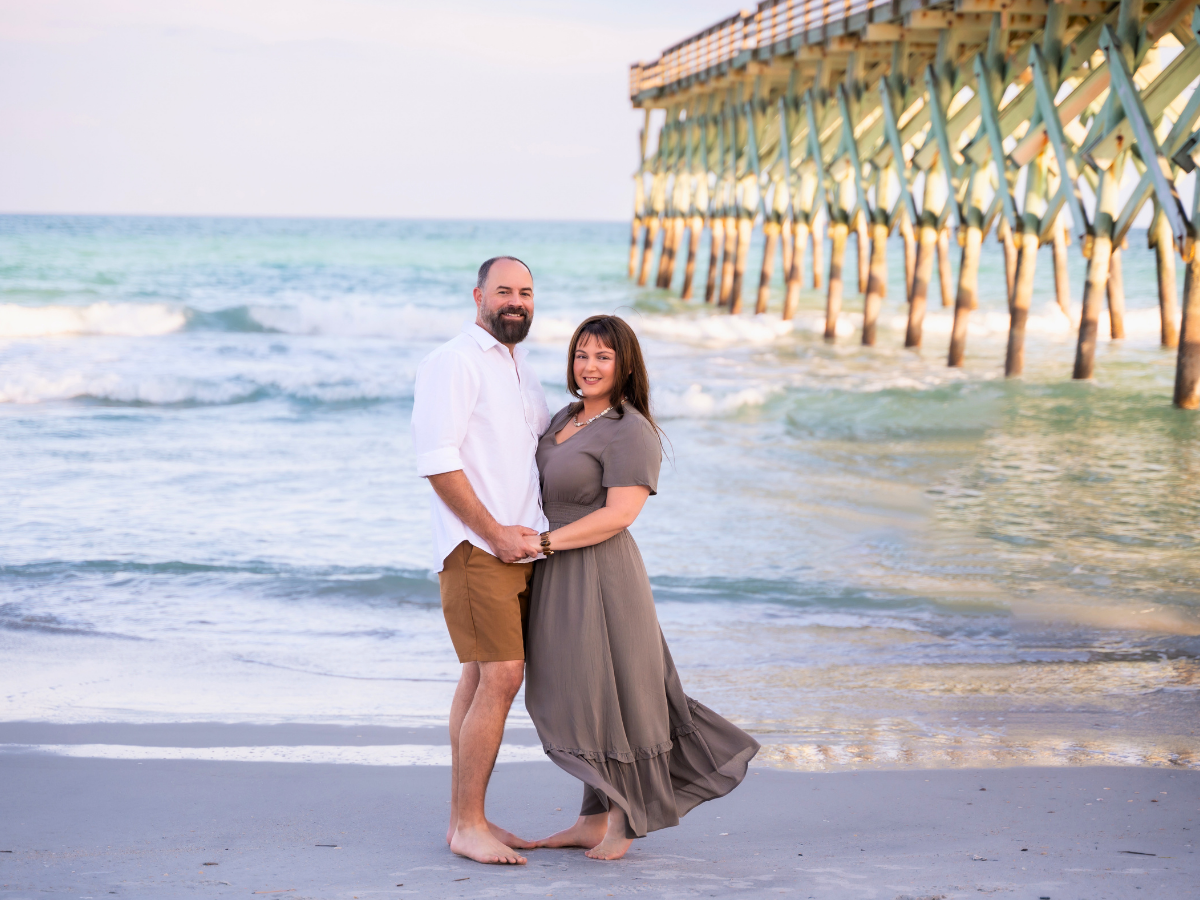 Couple standing barefoot on the beach, holding hands, with a wooden pier extending into the water behind them.