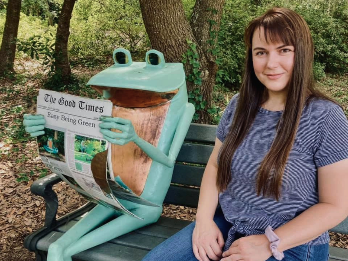 A turquoise frog statue on a park bench reads The Good Times newspaper beside a smiling woman in a striped shirt. The headline reads 'Easy Being Green.'