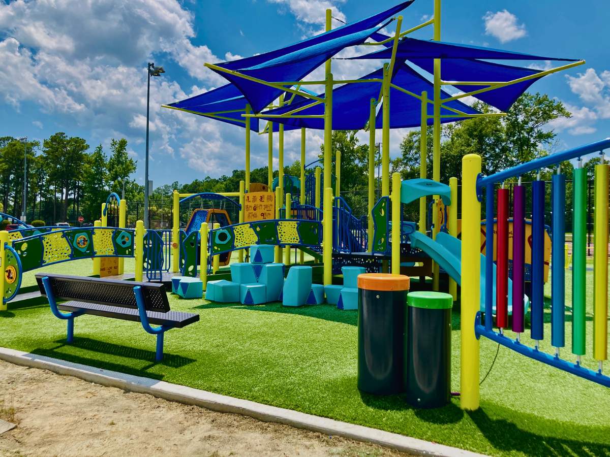 Colorful playground with blue shade canopies, climbing structures, slides, and a bench on artificial turf."