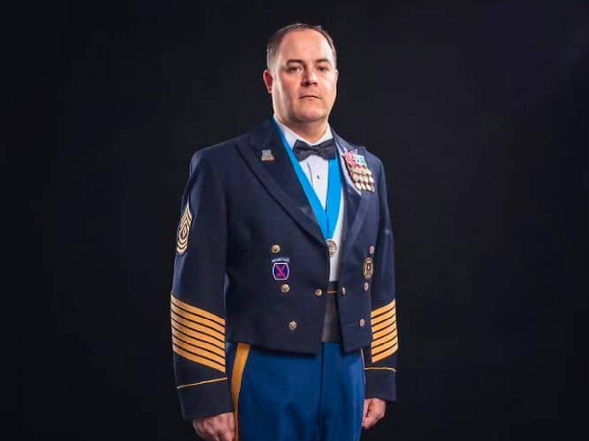 Man in formal military-style dress uniform with medals and blue ribbons, standing against a dark background.