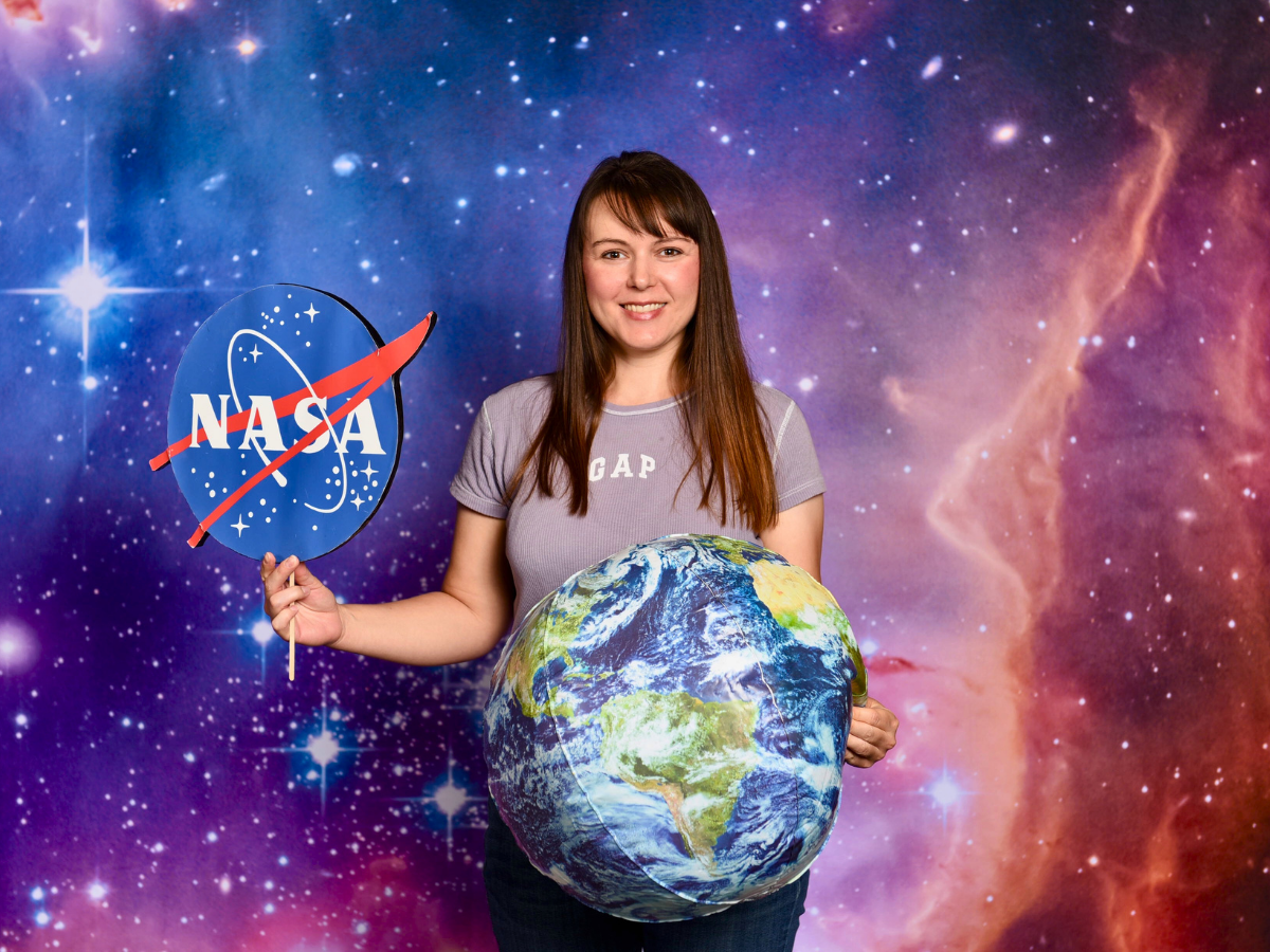 Smiling woman stands in a starry space backdrop, holding a NASA sign in one hand and a large inflatable Earth globe in front of her.