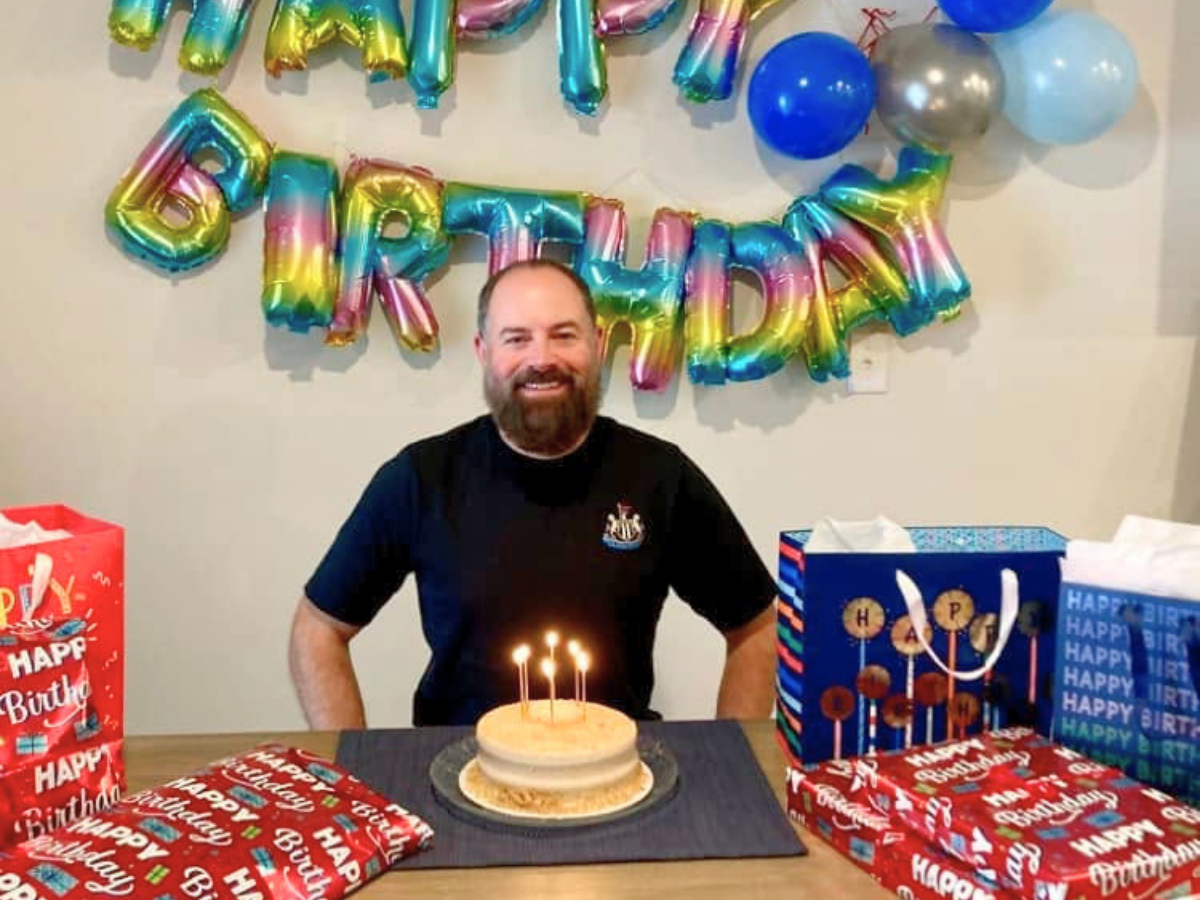 Man with a beard sitting at a table with a small cake and lit candles, birthday gifts and colorful balloons behind him.