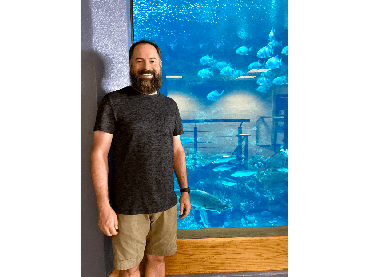Man with a beard stands in front of a large aquarium tank filled with blue water and fish, indoors.