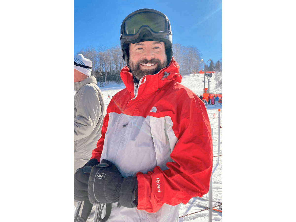 Smiling man in a red-and-gray ski jacket with goggles on a snowy ski slope, gloves on, sunny day.