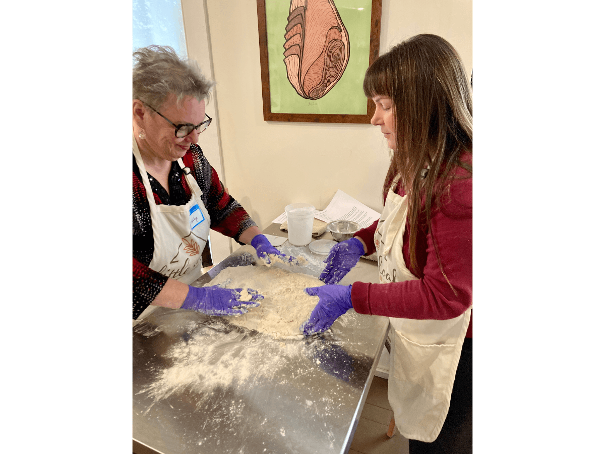 Two women wearing aprons and purple gloves kneading dough on a flour-dusted metal table, indoors as they bake together.