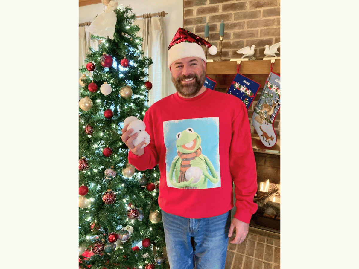 Smiling man in a Santa hat and red sweater with a frog image, holding a snowball beside a lit Christmas tree.