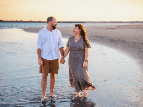 Couple holding hands and walking barefoot along the shoreline at sunset.