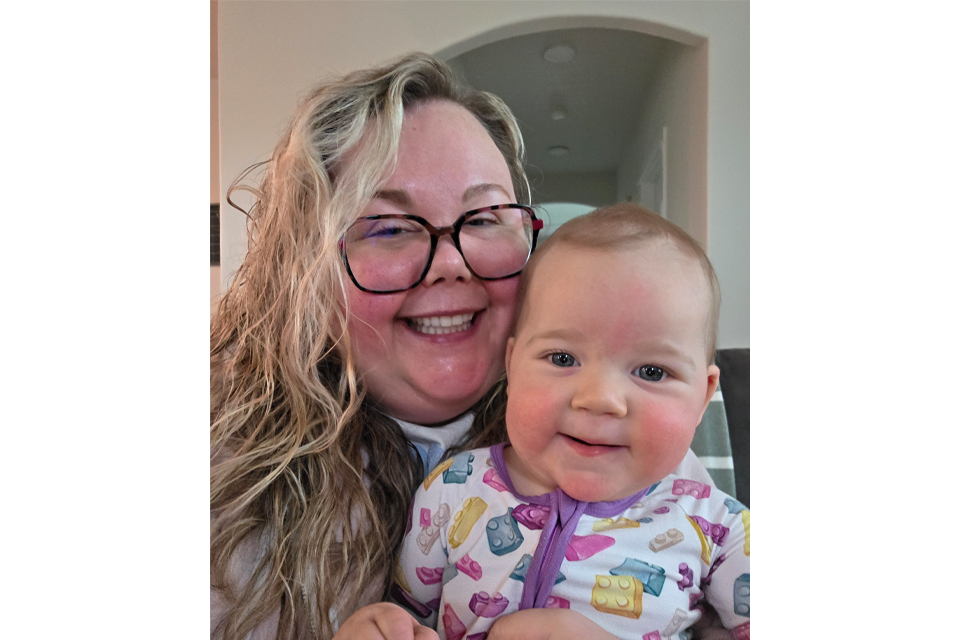 Mother with long wavy hair and red-framed glasses smiling with her baby, both looking at the camera indoors.