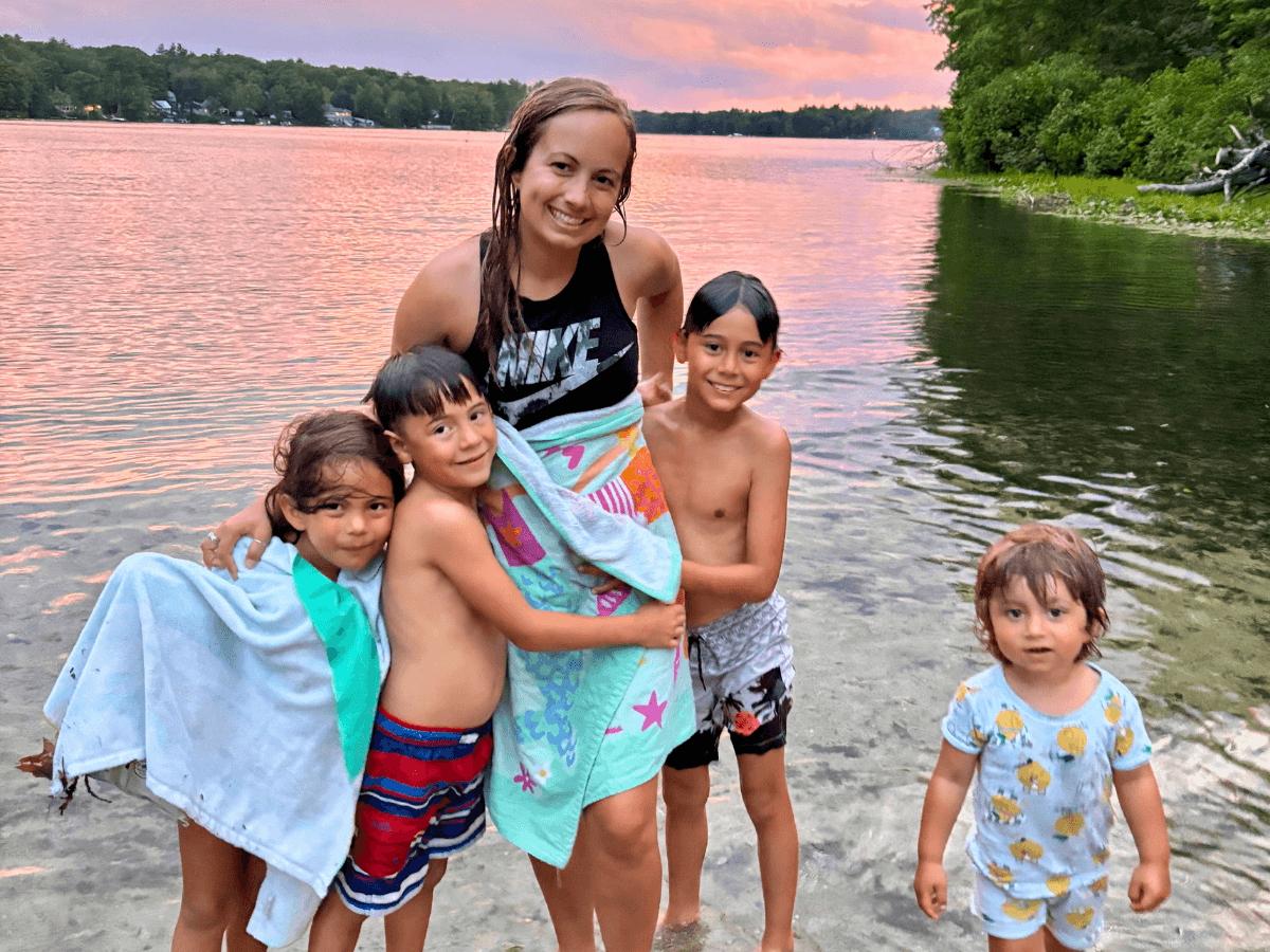 Woman and four children standing in shallow lake water at sunset, all smiling and wrapped in colorful towels, suggesting a family beach outing.
