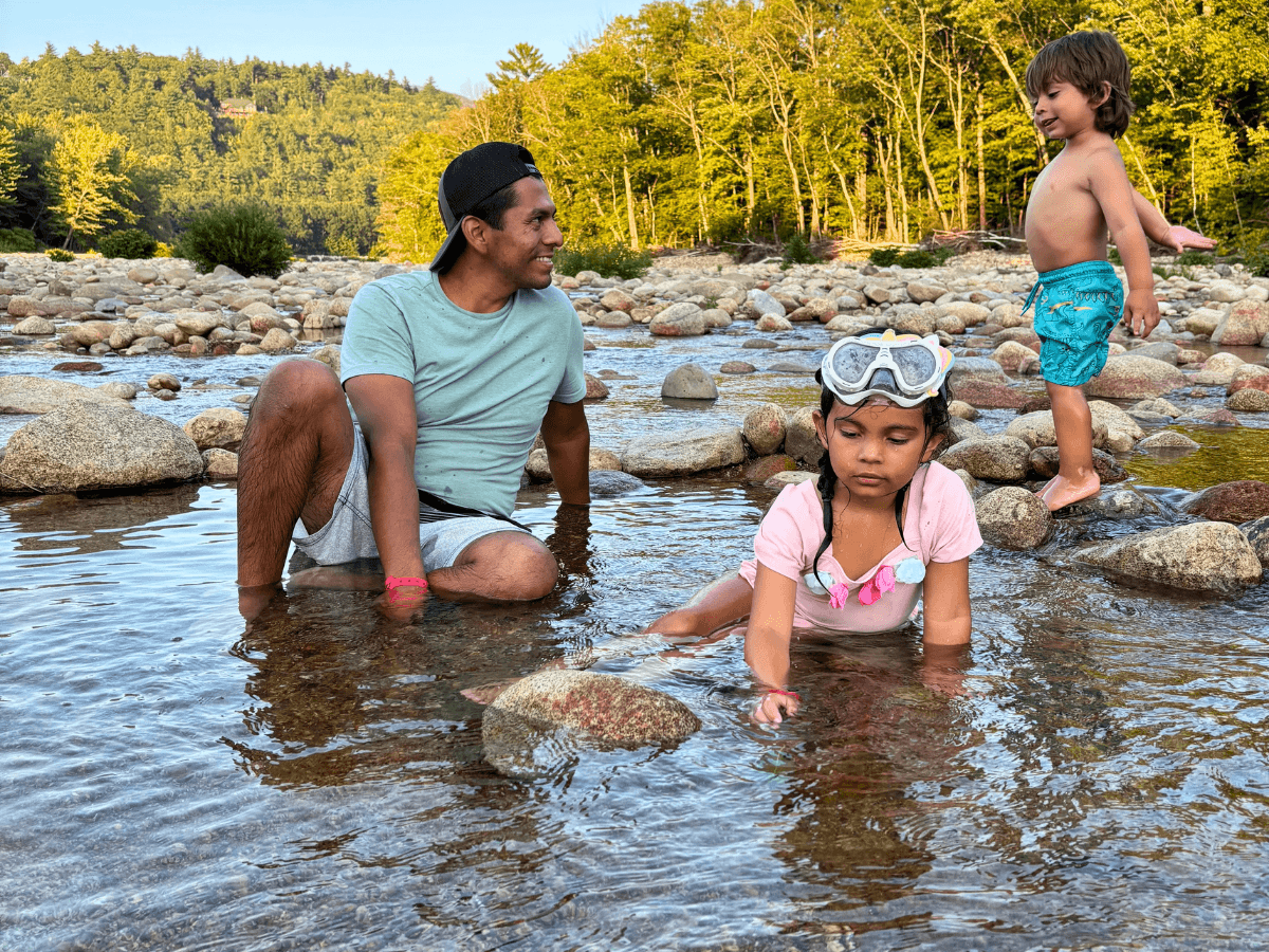 Man and two children wade in a shallow rocky river, girl wearing a snorkeling mask and pink top; a boy stands on rocks nearby.