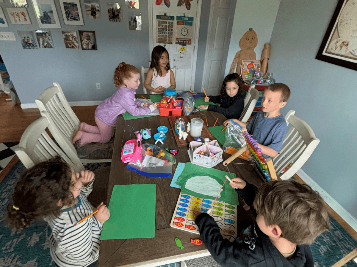 Group of young children seated around a wooden table doing crafts with green paper and colored pencils at home surveying supplies in a classroom-like setting.