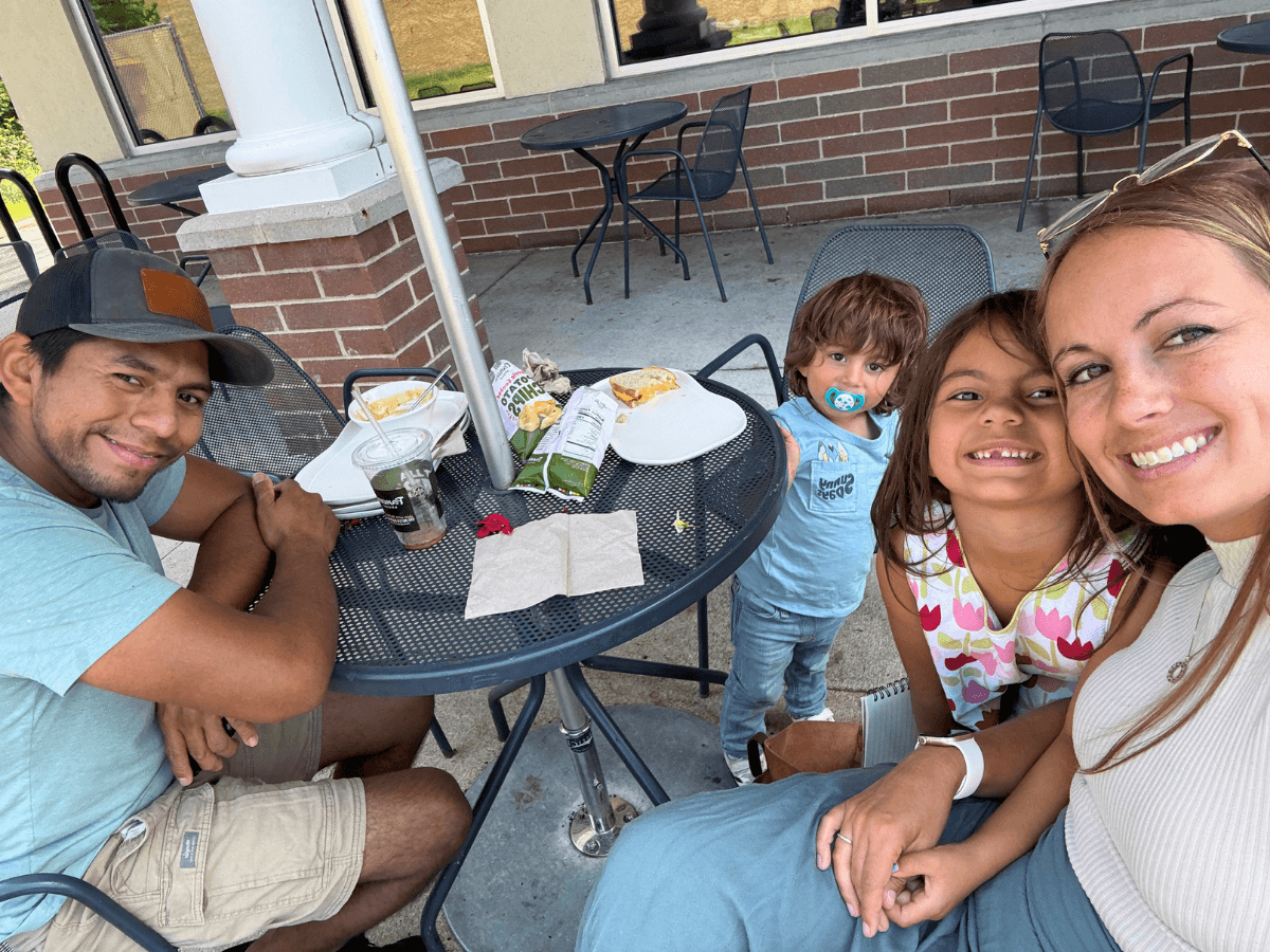A family of four sits at a round metal patio table outside, smiling at the camera with snacks and drinks on the table.