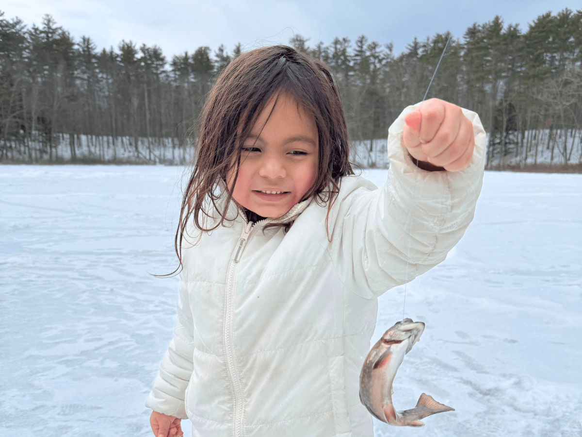 Young girl in a white winter jacket proudly holding up a small fish on a line over a frozen lake
