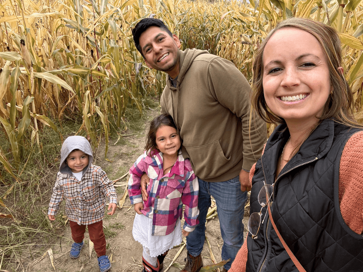 Woman takes a cheerful selfie with a man and two kids in a tall cornfield. All four smile at the camera in daytime light while wearing casual jackets and plaid shirts.