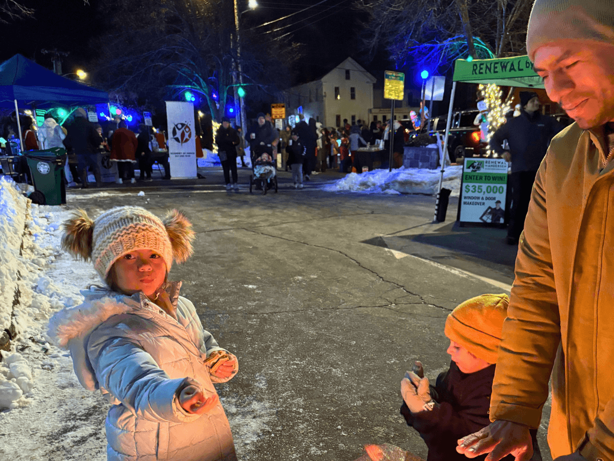 Young girl in a white coat and furry hat gestures toward the camera at a snowy outdoor festival. Nighttime winter market with colorful lights, tents, and people in the background; a man and child stand by a snowbank. Girl in a puffy coat and knit hat offers a snack at a winter festival, while a boy in an orange hat sits nearby. Crowd at a lit outdoor winter fair with stalls and signs; a smiling man stands beside two children. Children at a snowy street fair, girl reaching out toward the camera as a man and younger child observe.