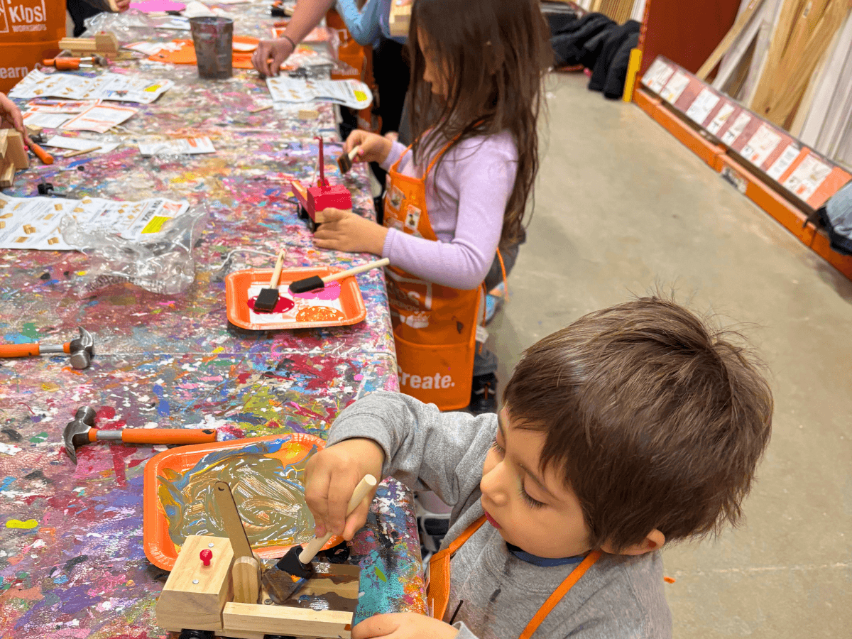 Children in orange aprons work on wooden crafts at a paint-splattered workshop table.