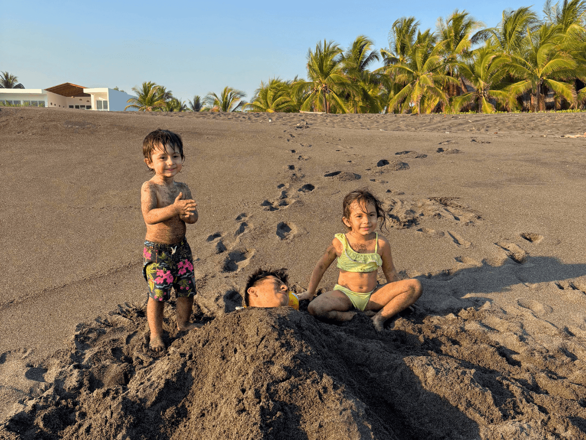 Three young children play on a sandy beach, one buried up to the shoulders in sand while two others sit and stand nearby, with palm trees and a modern house in the background.