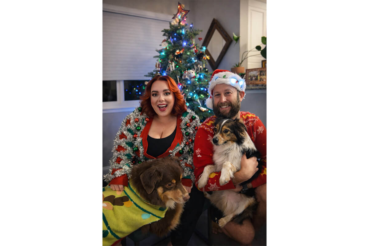 Smiling couple in festive sweaters posing with two dogs beside a lit Christmas tree indoors.