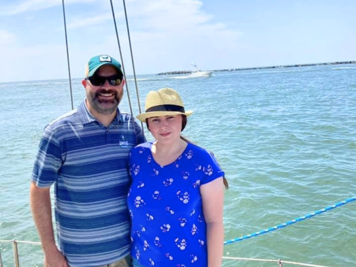 Two people posing on a boat near clear blue water, smiling at the camera; a man in a striped polo and a green cap next to a woman in a blue floral shirt and straw hat.