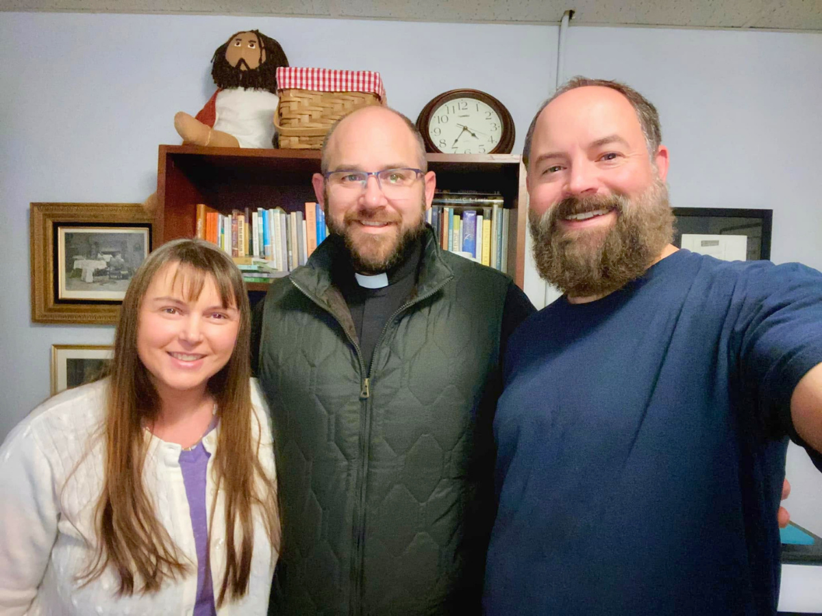 Three smiling adults pose for a selfie indoors, with a bookshelf, clock, and framed pictures in the background.