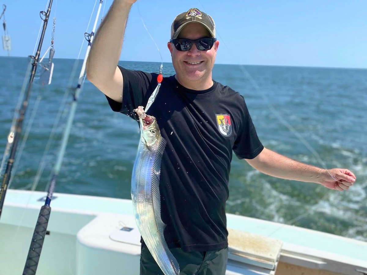 Man on a boat holding a long, silver fish he just caught, with the ocean in the background.