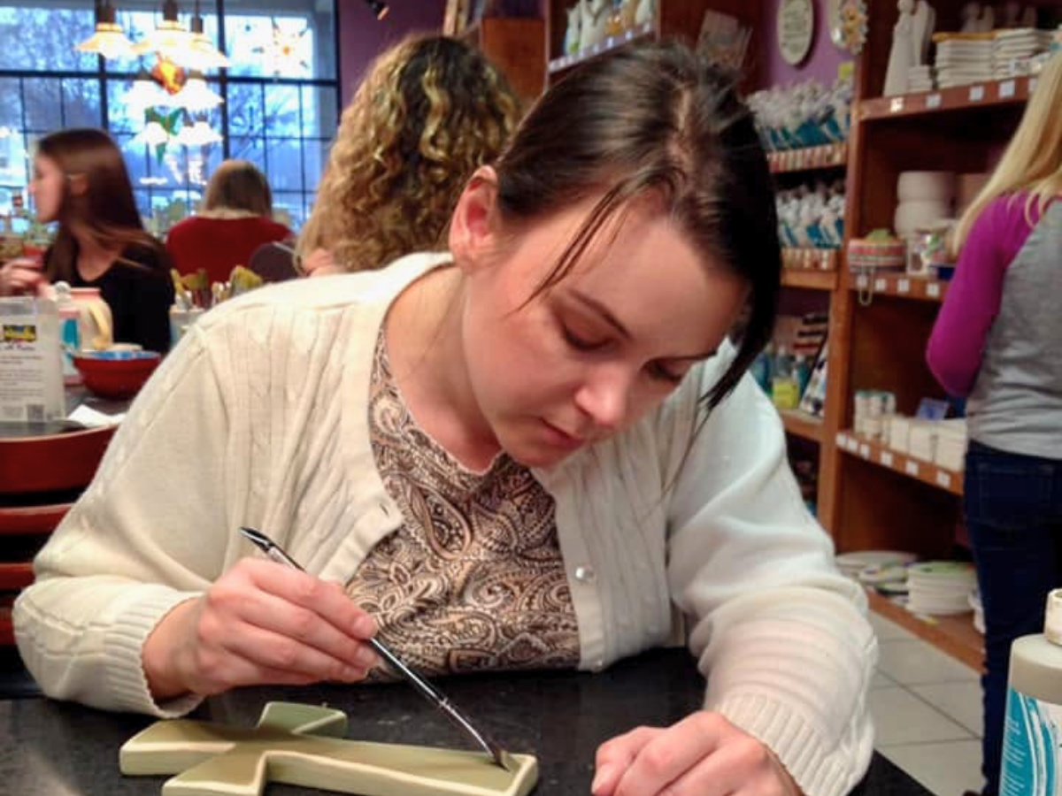 Woman leaning over a black table, painting a cross-shaped ceramic piece with a small brush in a craft store aisles filled with supplies behind her