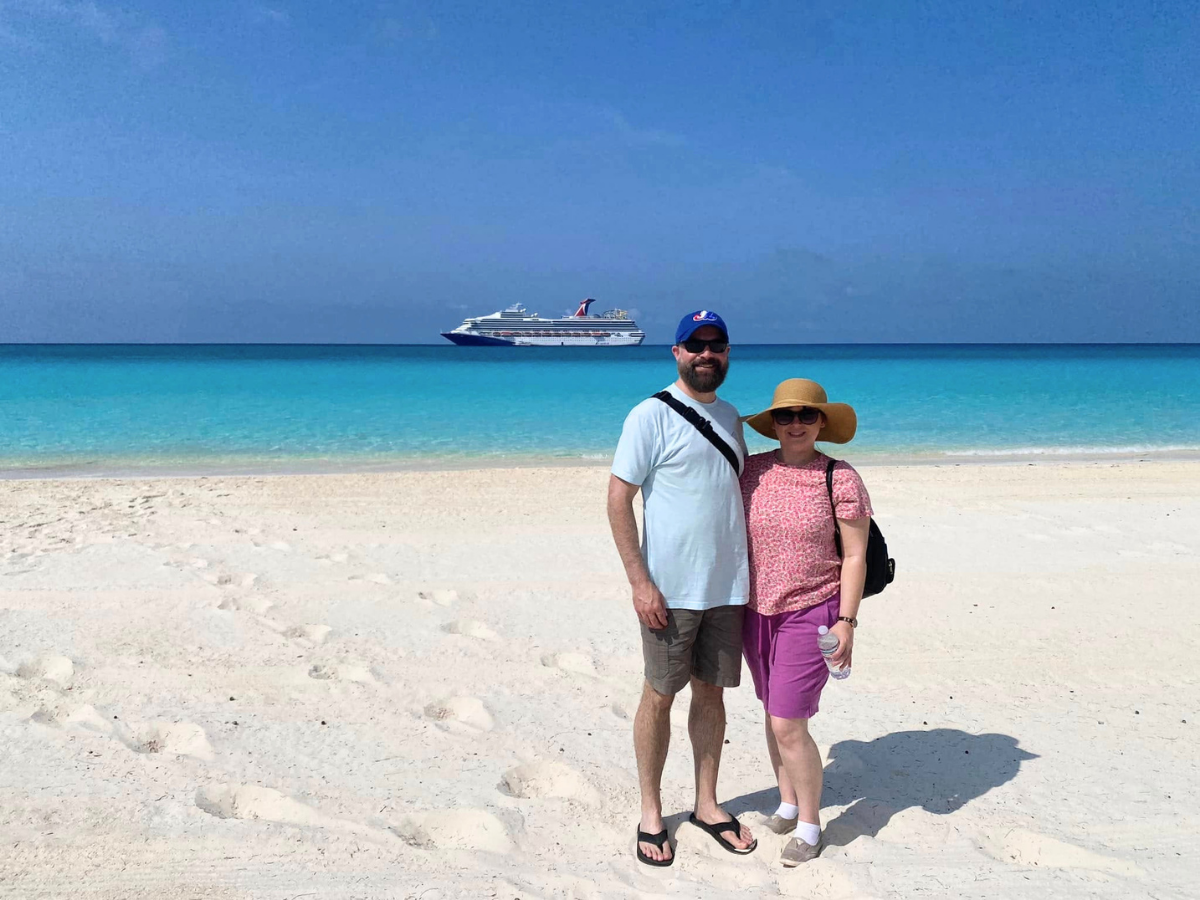 Couple standing on a white sandy beach with turquoise water and a cruise ship in the distance.