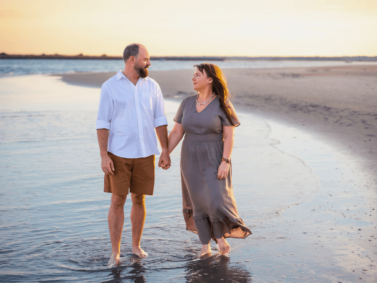 Couple walking hand in hand along the shoreline at sunset, barefoot in shallow water.