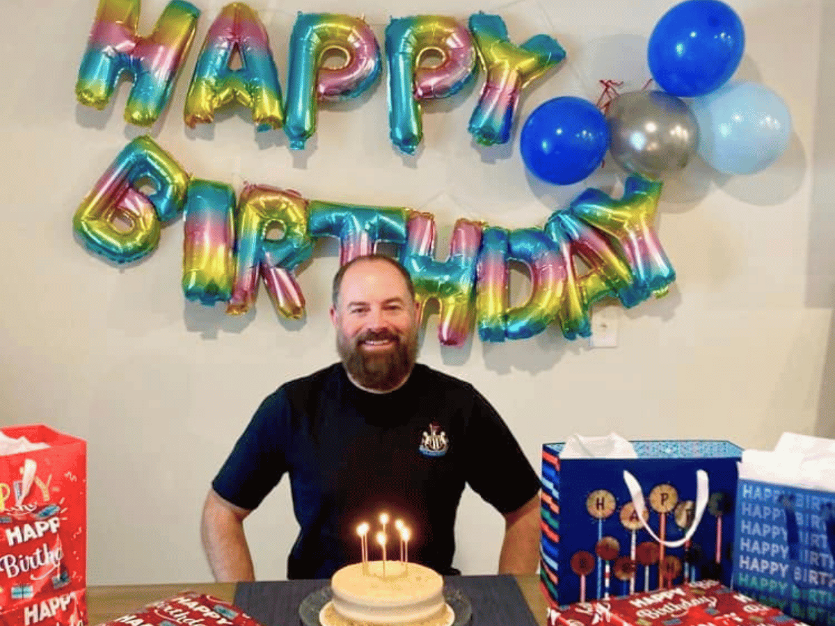 Smiling man at a birthday party with a small cake and lit candles, colorful 'Happy Birthday' balloons, and gift bags in the background.