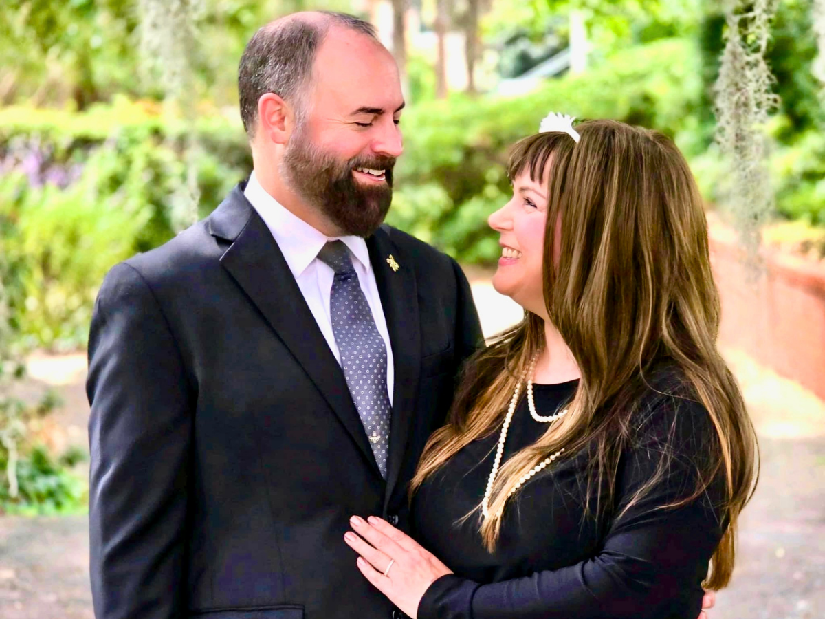 Smiling couple in formal attire standing close together in a lush garden.