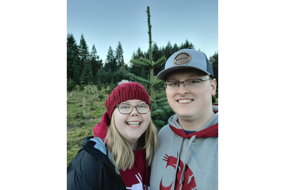 Smiling couple posing outdoors in a field of young evergreen trees, wearing hoodies and hats.