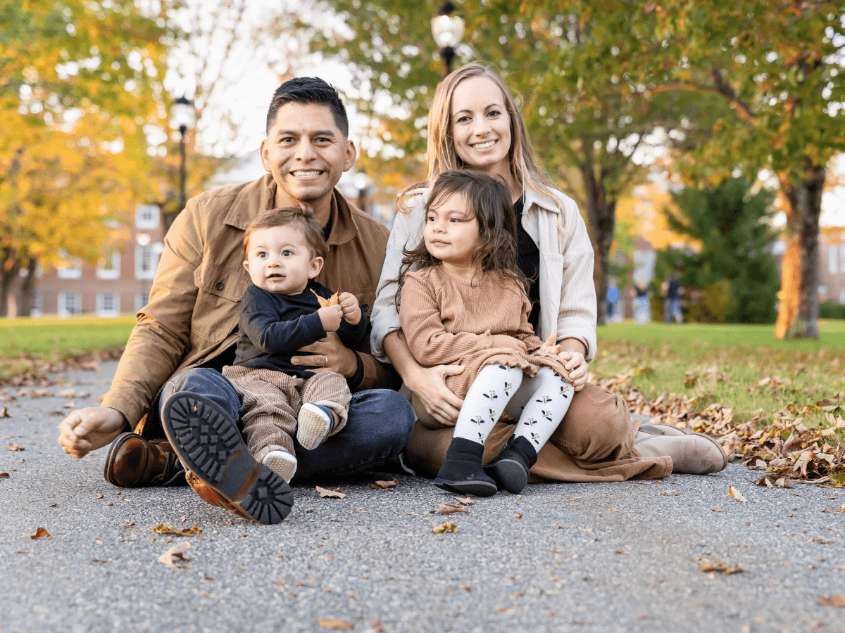 Family of four sitting on a park path covered in fallen leaves, smiling at the camera on an autumn day.