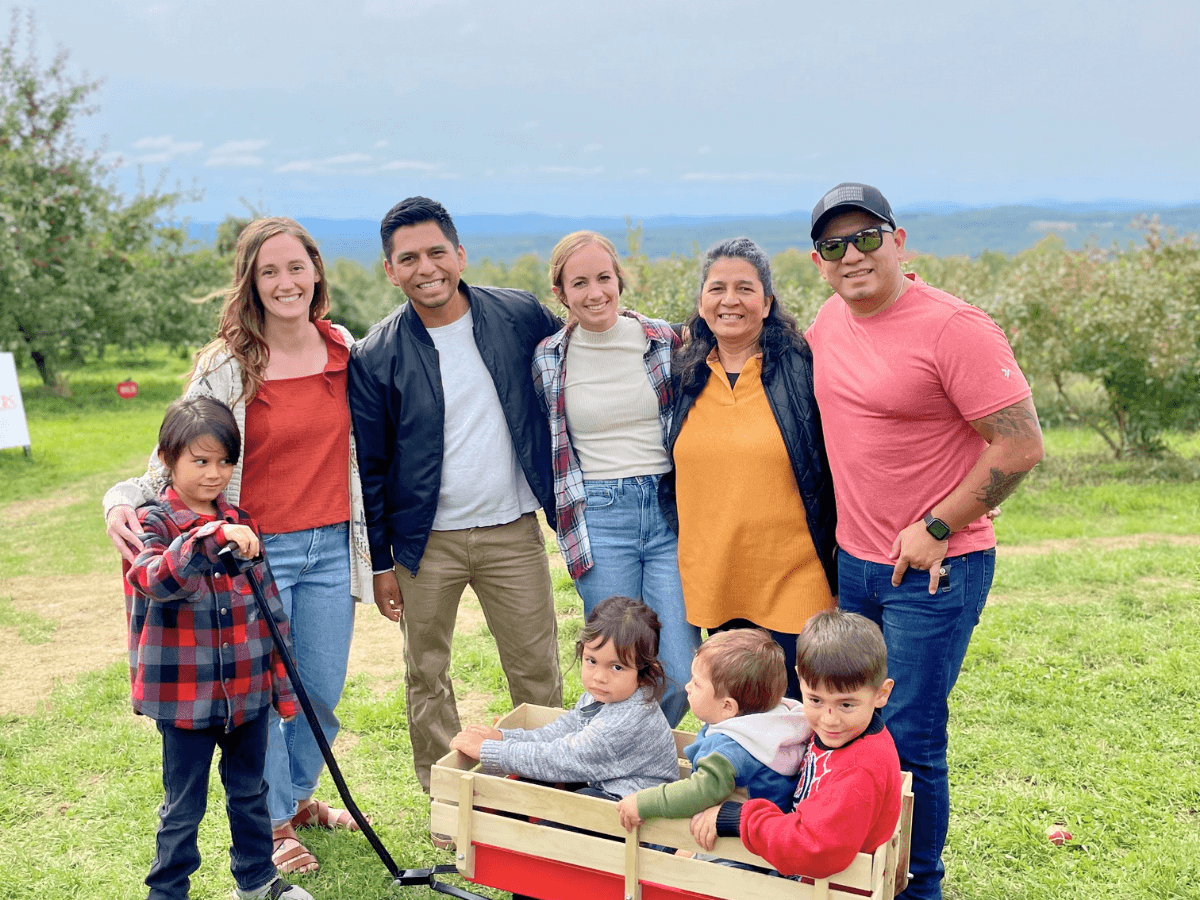 Group of adults and children posing together outdoors in a grassy field, with a wooden wagon of kids in front.