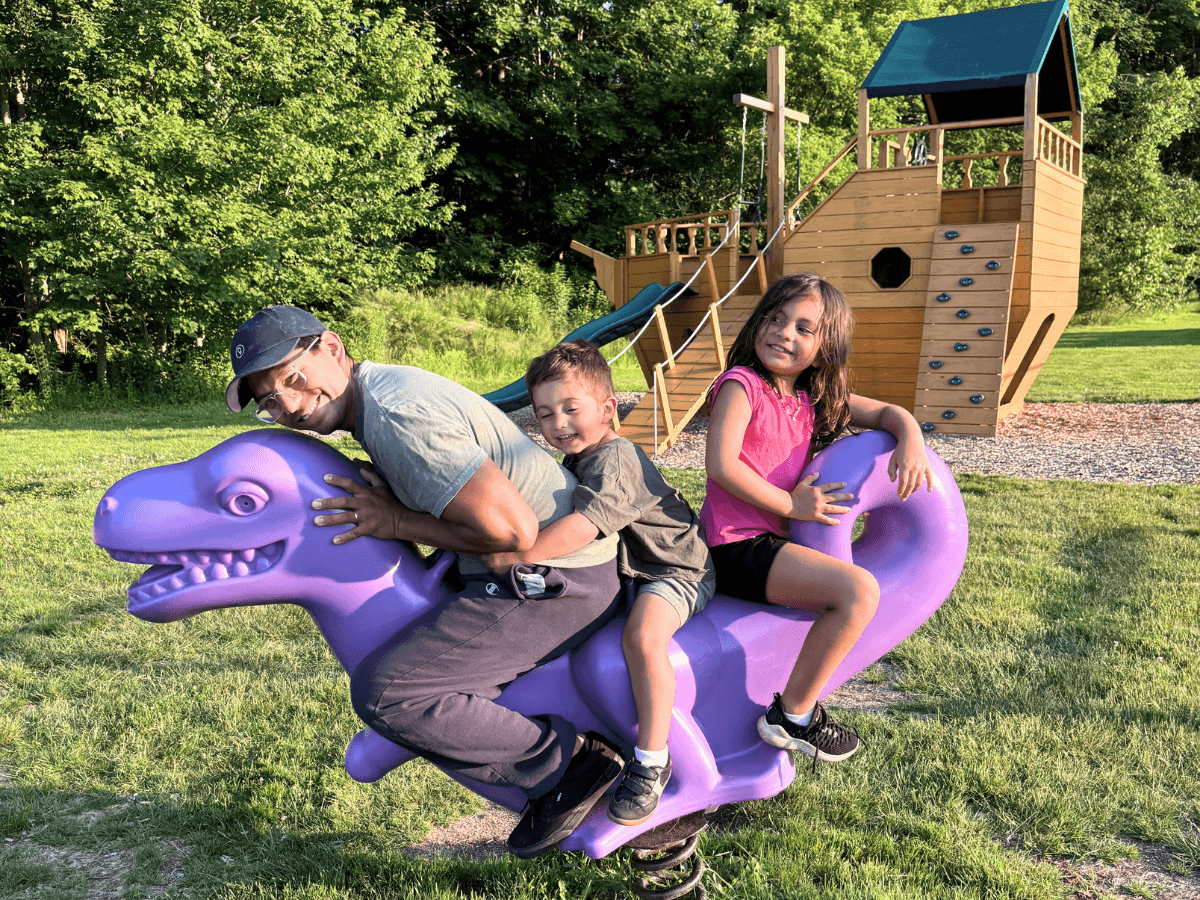 Man and two children ride a purple dinosaur playground spring rider in a sunny park with a wooden climbing structure in the background.