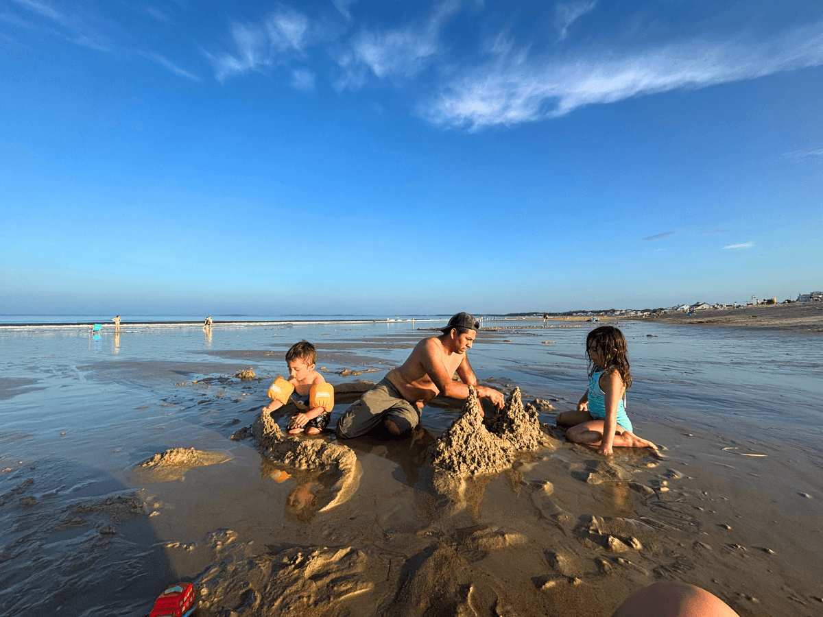Group of three at the shoreline building sandy castles on a sunny beach with calm water and blue sky behind them