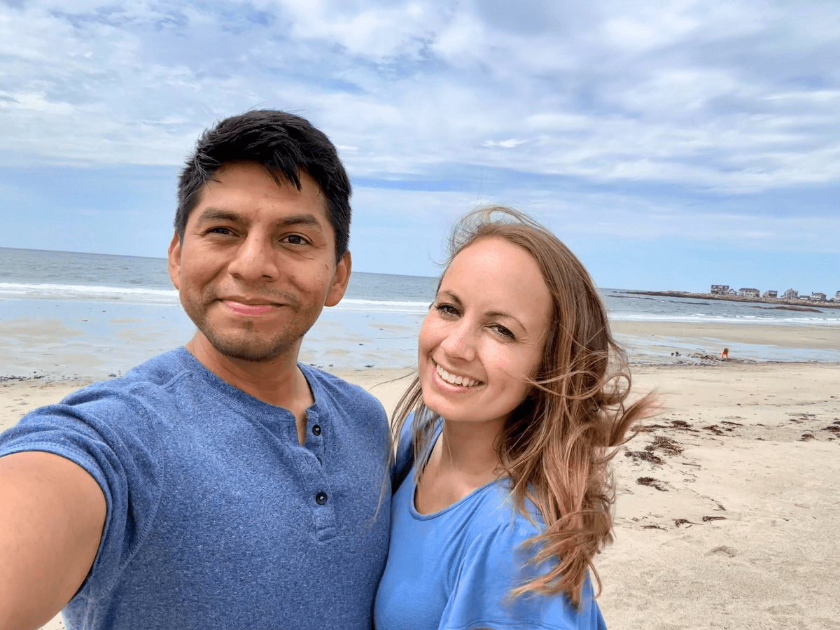 Couple taking a selfie on a sandy beach with the ocean and cloudy sky behind them, smiling at the camera.
