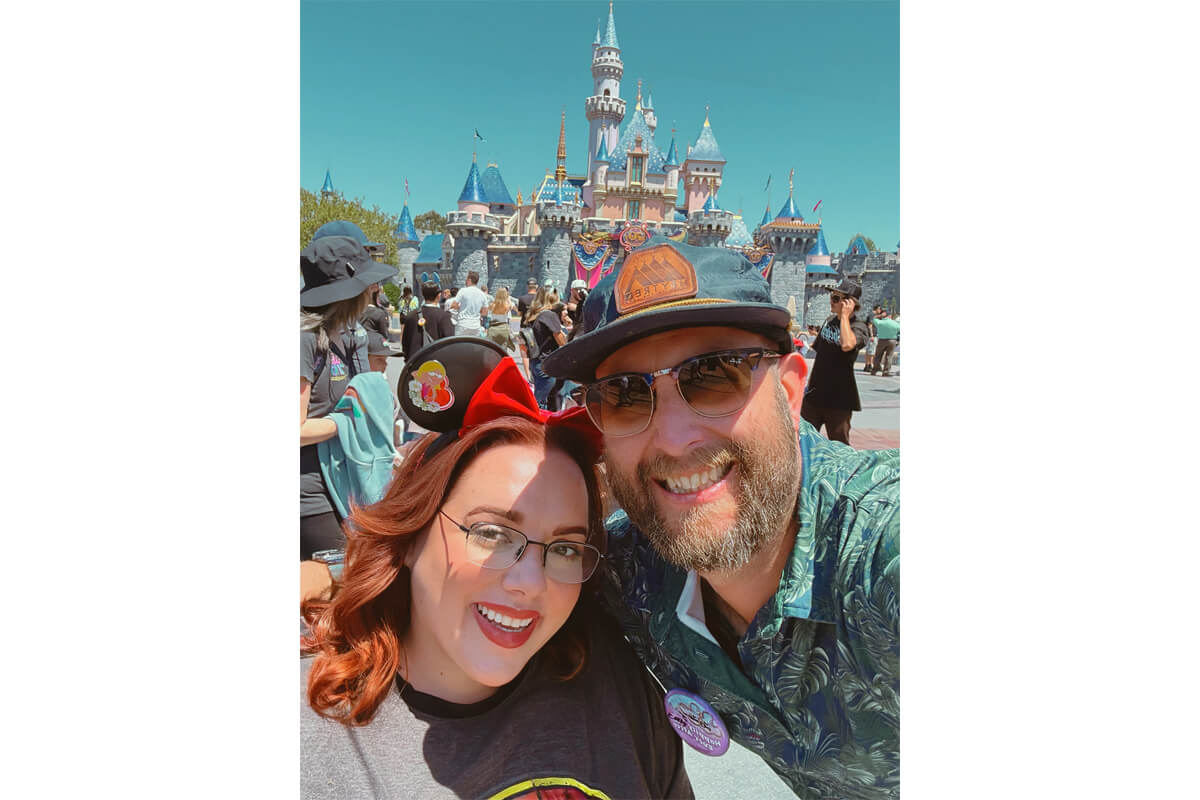Smiling couple taking a selfie at a sunny Disney park with a castle in the background; woman wears Minnie ears, man wears sunglasses and a cap.