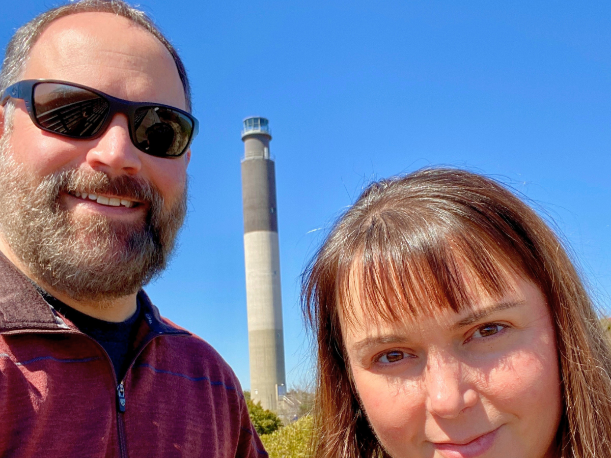 Smiling man wearing sunglasses and a beard poses for a selfie with a woman, a tall lighthouse behind them against a blue sky.