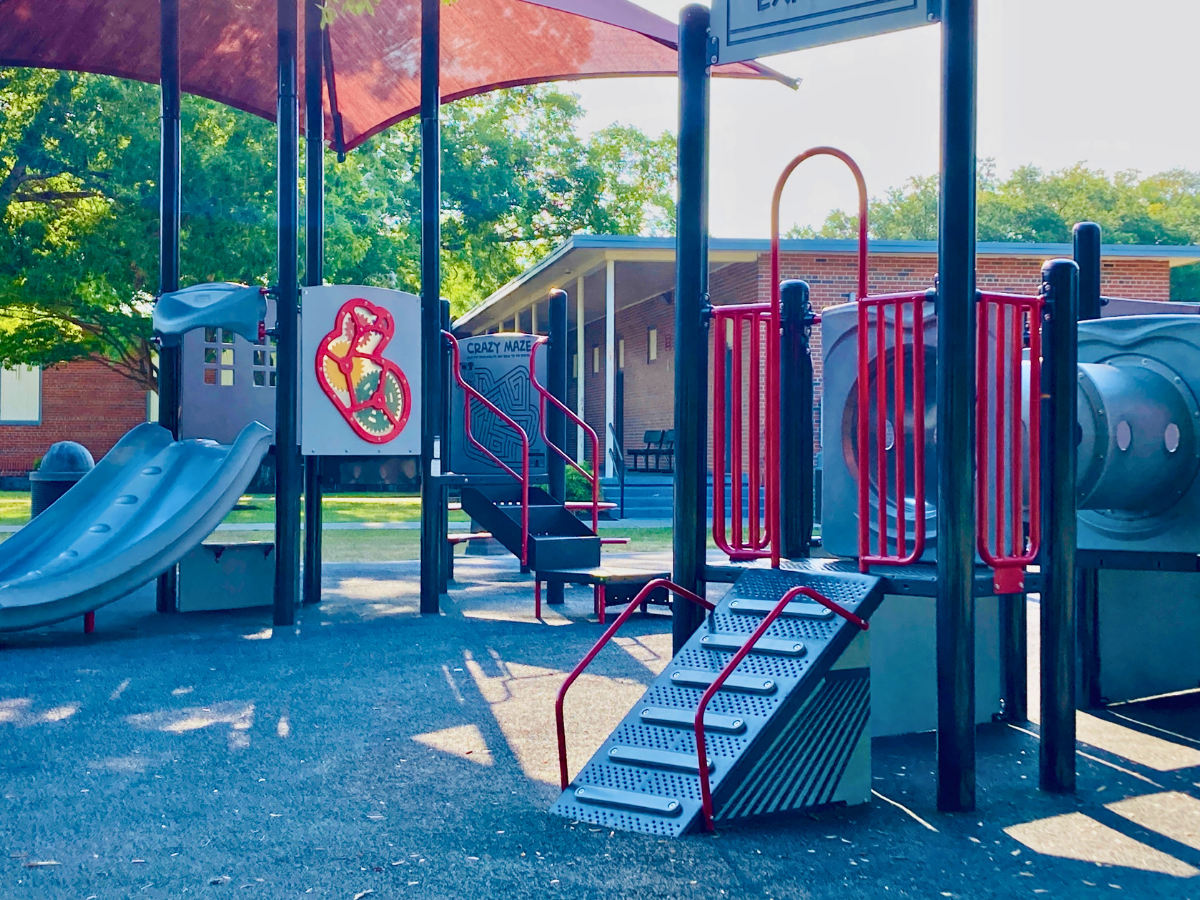 Colorful playground with slides, stairs, and red railings under a shade canopy; a brick building is in the background.