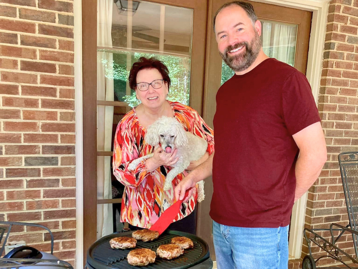 Woman and man grill burgers on a patio while holding a small white dog; smiling for a photo.