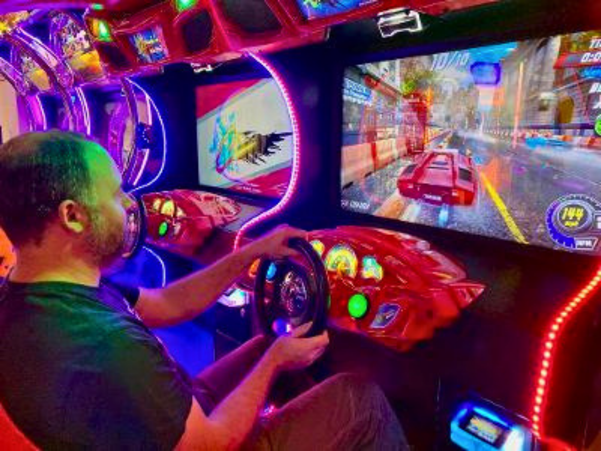 Man driving a neon-lit arcade racing simulator, gripping a red steering wheel while a high-speed car game runs on a large screen.
