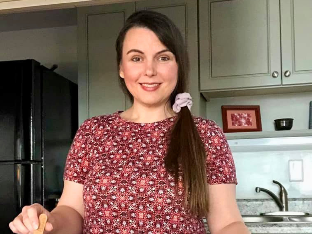 Smiling woman with long dark hair in a red floral shirt standing in a kitchen, holding a small cookie.