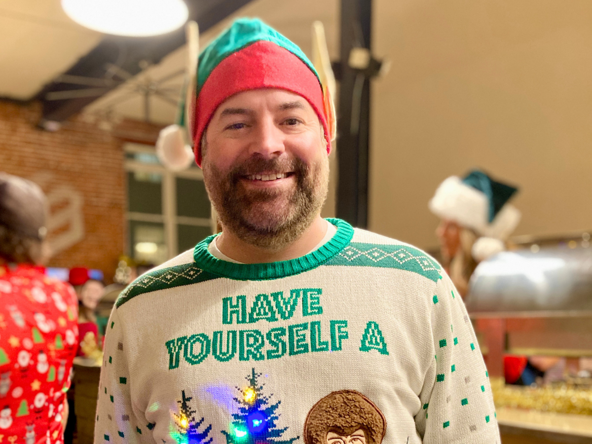 Smiling man in a festive sweater with 'HAVE YOURSELF A' text and a red-green elf hat at a holiday party.