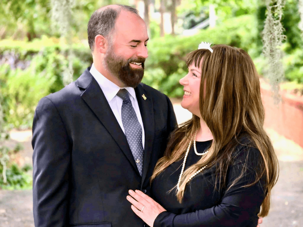 Smiling couple in formal attire posing outdoors, man in suit and tie, woman in black dress with pearls and headband.