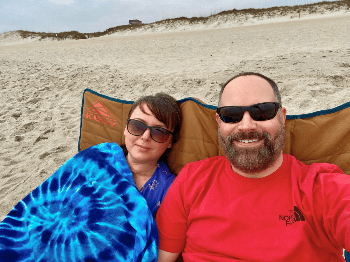 Smiling couple wearing sunglasses on a sandy beach, lounging in a tan beach chair with a blue tie-dye blanket.