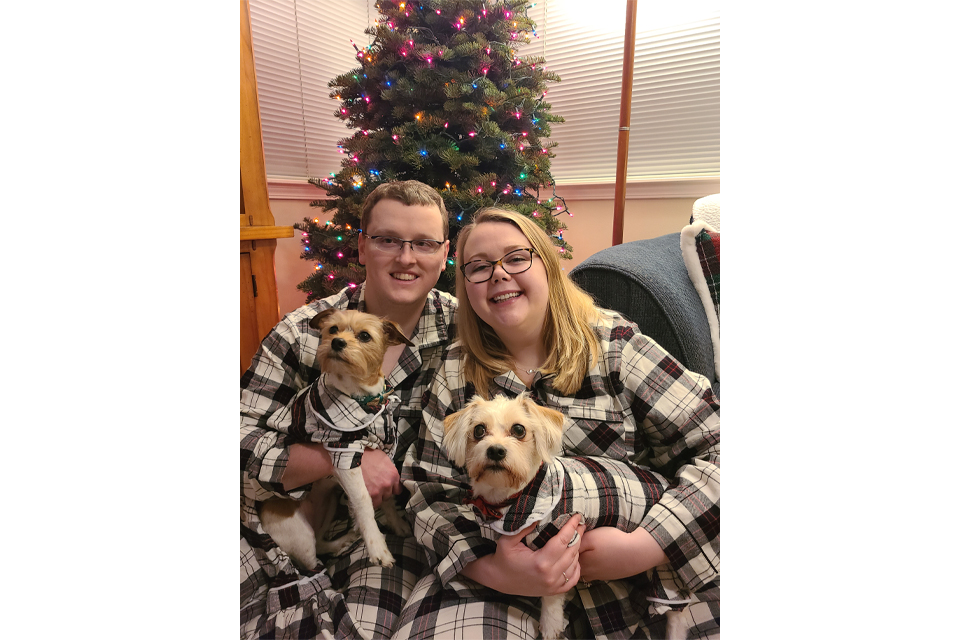 Couple wearing plaid shirts holding two small dogs in front of a Christmas tree with colorful lights.