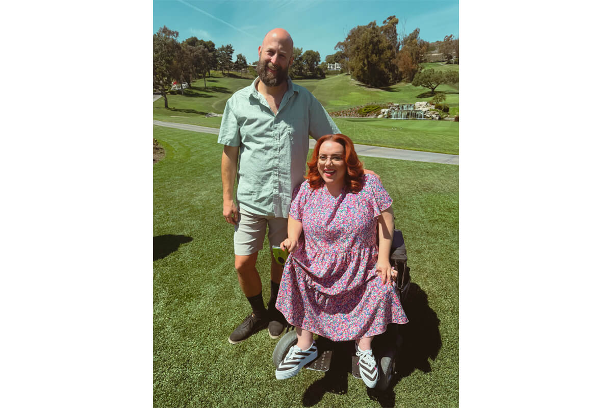 Man with a beard in a green shirt stands beside a smiling woman in a floral dress seated in a wheelchair on a sunny golf course, with trees and a fountain in the background.