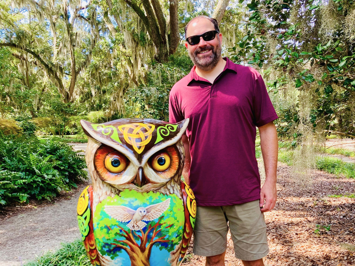 Man wearing a maroon polo and sunglasses stands beside a brightly painted owl sculpture in a sunny park.