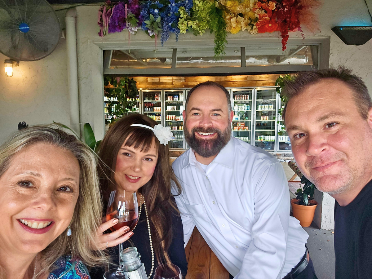 Four friends taking a selfie at an outdoor bar, smiling and holding drinks, with colorful flowers above.